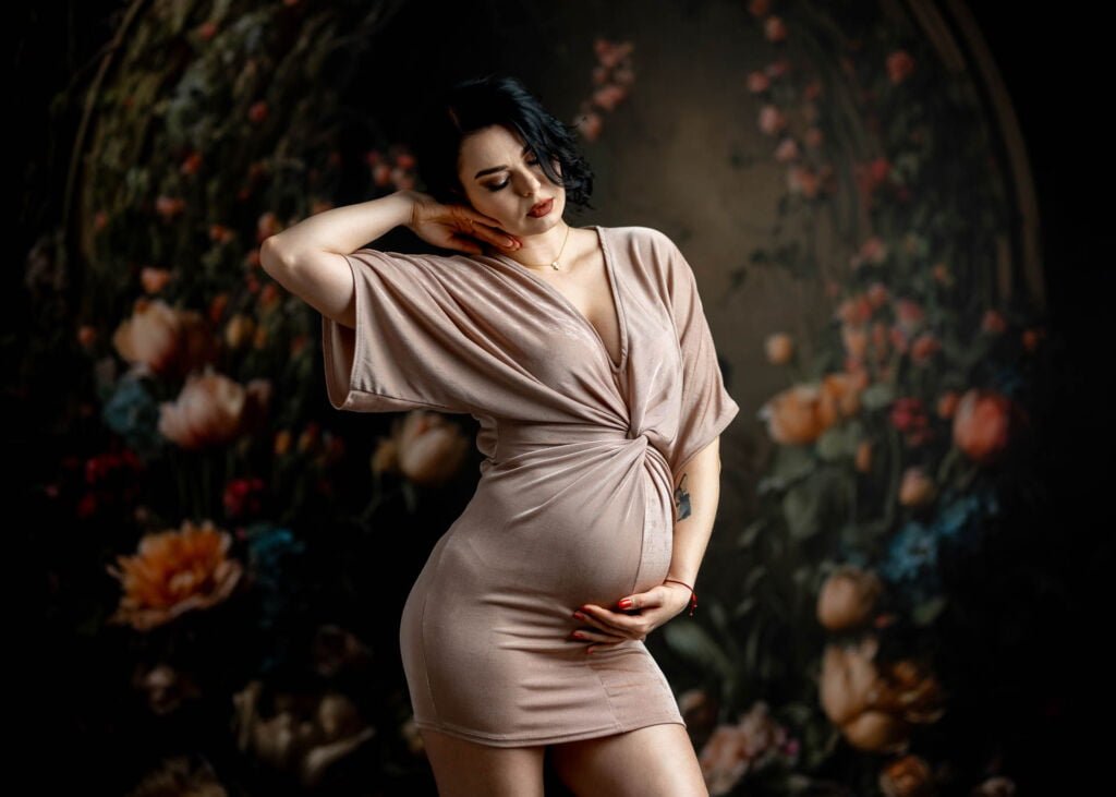 Przyszła mama w eleganckiej, białej sukni ciążowej, pozuje w studio fotograficznym w Słupsku, otoczona delikatnymi tkaninami tworzącymi romantyczną atmosferę